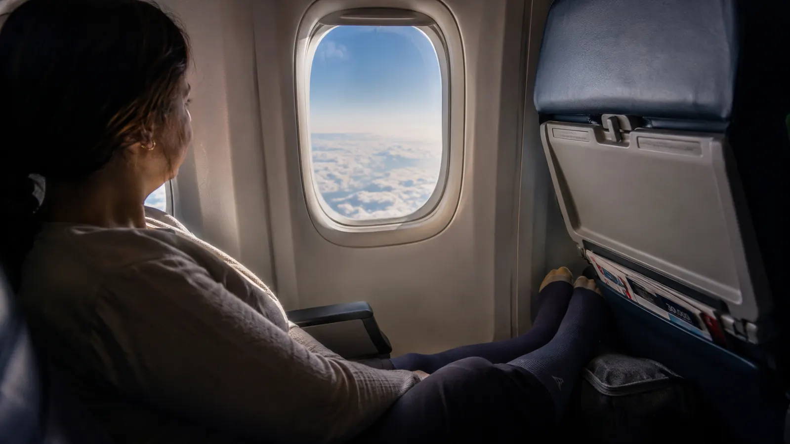 Traveler in window seat on a long-haul flight looking out at clouds, feet resting on carry-on under the seat ahead