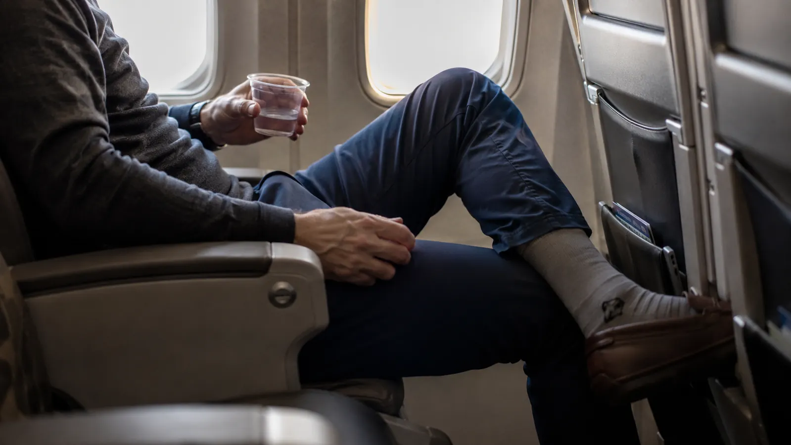 Traveler in compression socks settling into a window seat on a long-haul flight, carry-on bag overhead