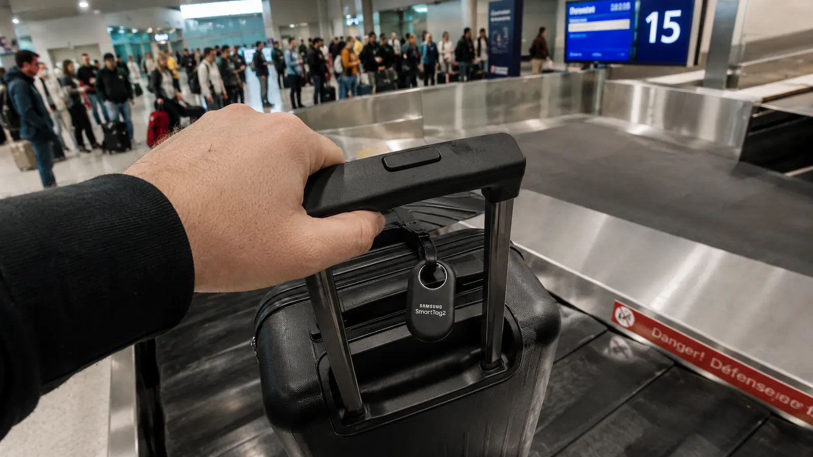 Samsung SmartTag2 clipped to a luggage handle on a busy airport conveyor belt